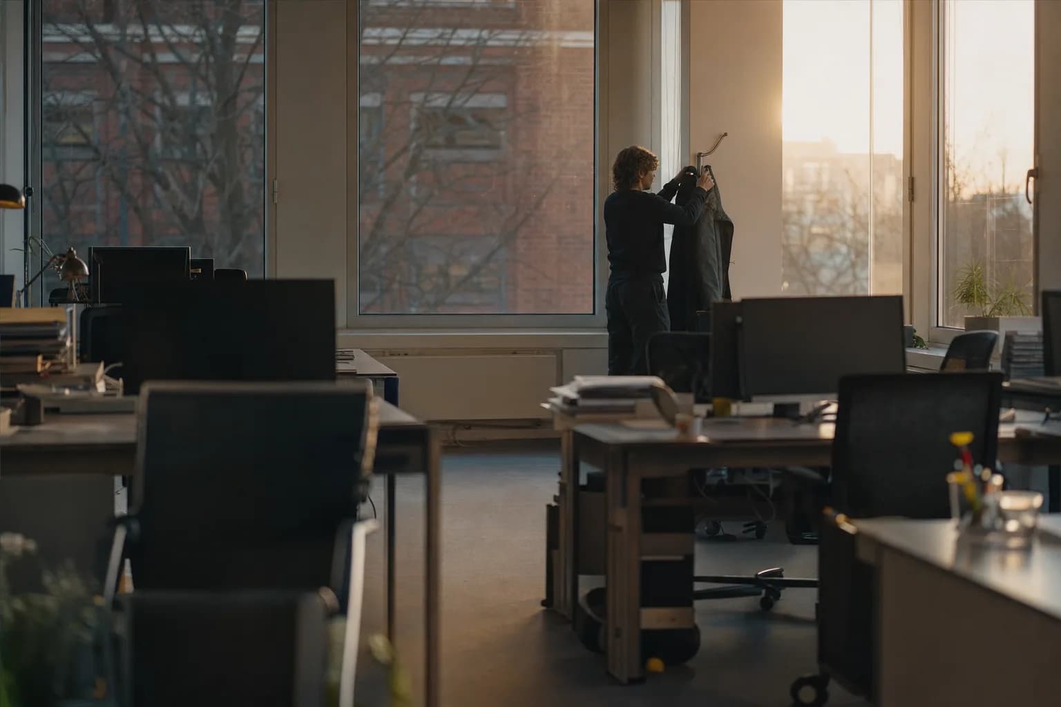 An office at eight in the morning, empty desks with monitors off, one person hanging a coat by the window