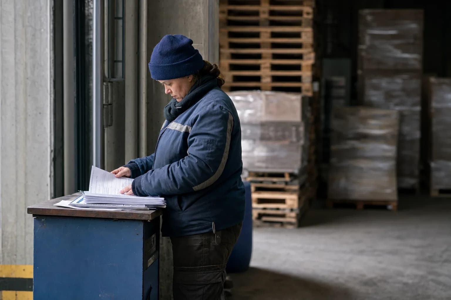A worker at a standing desk by a warehouse dock, going through a stack of paperwork, wooden pallets soft in the background